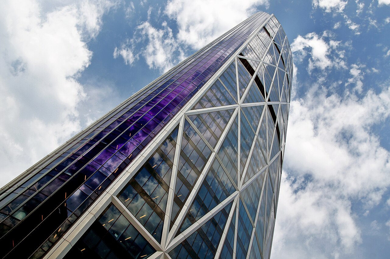 Looking straight up the glass and steel facade of The Bow tower in Calgary against a blue sky with clouds